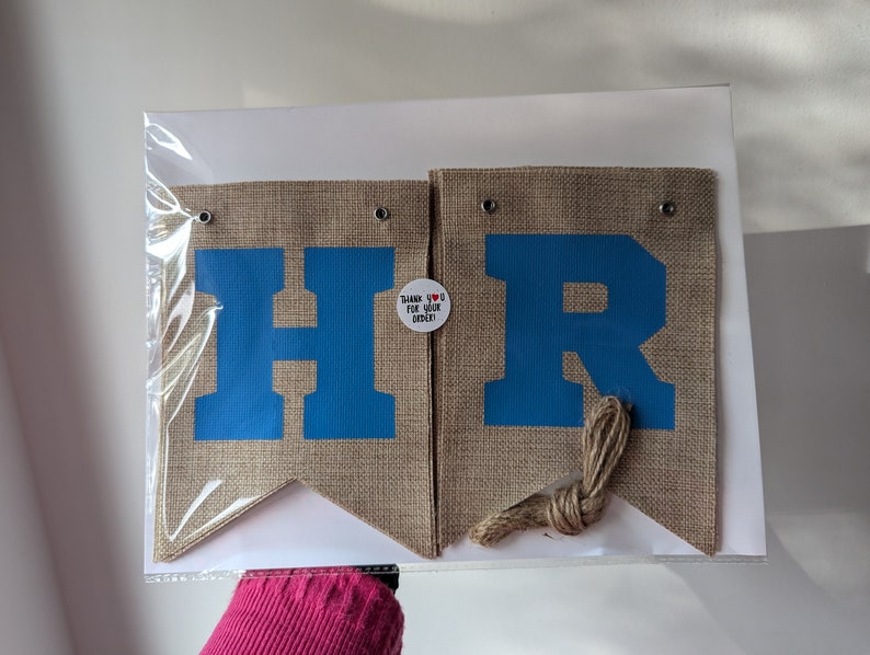 Puede incluir: Una pancarta de arpillera con las letras "HR" en azul sobre un fondo beige. La pancarta est&aacute; unida con un cordel y tiene una peque&ntilde;a etiqueta que dice "&iexcl;Gracias por tu pedido!" con un coraz&oacute;n rojo.