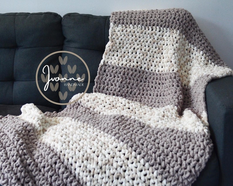May include: A gray and white striped crocheted throw blanket draped over a gray couch. The blanket is made with thick yarn and has a chunky texture. The blanket is handmade and has a tag that says "Yvonne Handmade".