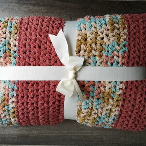 May include: A crocheted throw blanket with a multi-colored pattern in shades of pink, blue, beige, and brown. The blanket is tied with a white ribbon.