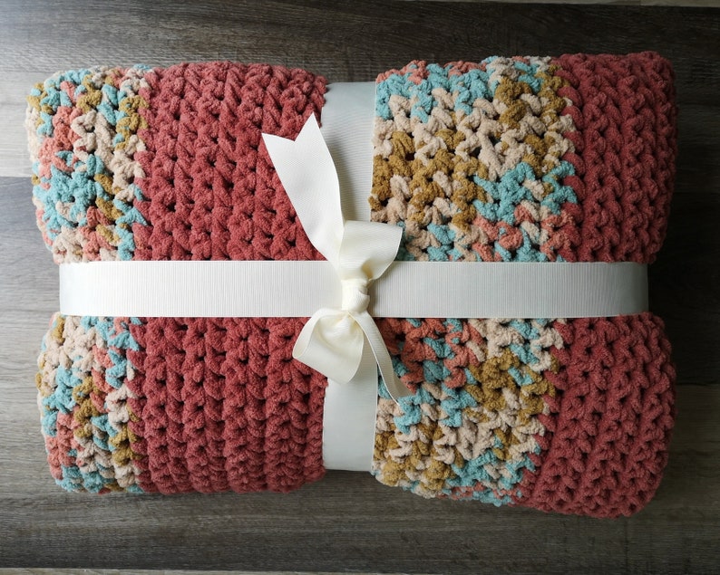May include: A crocheted throw blanket with a multi-colored pattern in shades of pink, blue, beige, and brown. The blanket is tied with a white ribbon.