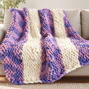 Könnte beinhalten: Eine grob gestrickte Wohndecke mit abwechselnden vertikalen Streifen aus cremefarbenem, hellviolettem und dunkelviolettem Garn. Die Decke ist mit einer dicken, geschlungenen Textur gefertigt und über ein Sofa in neutraler Farbe drapiert.