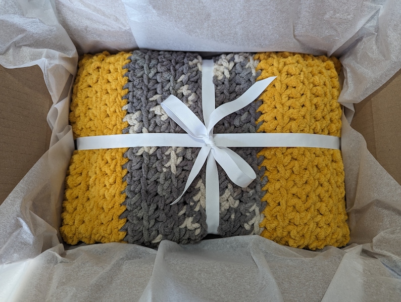 May include: A yellow and gray crocheted blanket tied with a white ribbon, packaged in a white tissue-lined box.