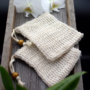 Könnte beinhalten: Zwei beige gewebte Seifensäckchen mit Kordelverschlüssen und Holzperlen, die auf einem verwitterten Holztablett liegen. Weiße Orchideenblüten sind im Hintergrund, mit grünen Blättern im Vordergrund. Die Beutel bestehen aus Naturfaser.