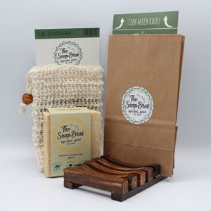 Può includere: Un sacchetto di carta marrone con un adesivo verde che dice "ZZEH MICH RAUS!" Il sacchetto è accanto a un porta sapone di legno con una saponetta all'interno. Il sapone è etichettato "The Soap Bros Natural Soap". C'è un sacchetto di rete bianco con una perla di legno in cima.