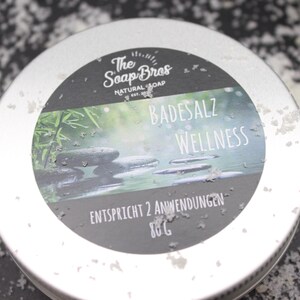 Könnte beinhalten: Eine silberne Dose mit einem schwarz-weißen Etikett, auf dem "The Soap Bros Natural Soap" und "Badesalz Wellness" steht, sowie eine grüne Bambuspflanze und graue Steine auf dem Etikett. Das Etikett trägt außerdem die Aufschrift "Entspricht 2 Anwendungen 80g".