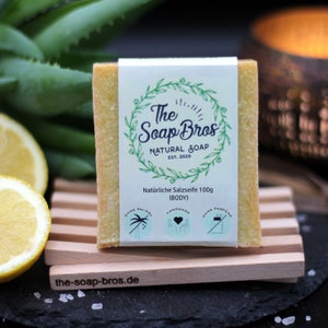 Könnte beinhalten: Eine natürliche Salzseife mit einem grün-weißen Etikett, auf dem "The Soap Bros Natural Soap Est. 2020" steht. Das Etikett enthält auch den Text "Natürliche Salzseife 100g (BODY)" und drei Symbole: eine Palme, ein Herz und eine Flasche mit Deckel. Die Seife liegt auf einem Holzseifenhalter.