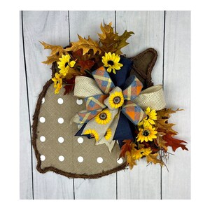 May include: A brown burlap acorn-shaped door hanger with white polka dots. A blue and brown plaid bow with a tan ribbon is tied in the center. The bow is decorated with small yellow sunflowers. The hanger is surrounded by brown and orange fall leaves.