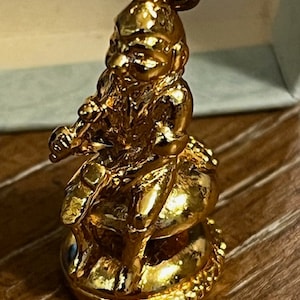 Può includere: Un ciondolo dorato a forma di gnomo seduto su un fungo. Lo gnomo indossa un cappello a punta e ha la barba. Il ciondolo ha un anello in cima per essere attaccato a una collana o un braccialetto.