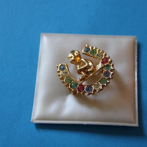 K&ouml;nnte beinhalten: Eine goldfarbene Brosche mit einer kleinen Figur, die auf einem hufeisenf&ouml;rmigen Sockel sitzt. Das Hufeisen ist mit bunten Strasssteinen verziert.