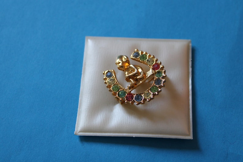 K&ouml;nnte beinhalten: Eine goldfarbene Brosche mit einer kleinen Figur, die auf einem hufeisenf&ouml;rmigen Sockel sitzt. Das Hufeisen ist mit bunten Strasssteinen verziert.