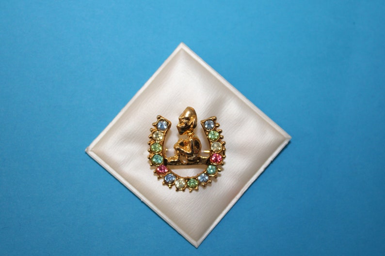 K&ouml;nnte beinhalten: Eine goldene Hufeisen-Brosche mit einer kleinen Figur eines sitzenden Menschen im Hufeisen. Das Hufeisen ist mit bunten Strasssteinen verziert.