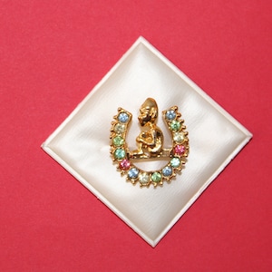 Puede incluir: Un amuleto de herradura de oro con una pequeña figura sentada en la parte superior. La herradura está decorada con brillantes de colores.