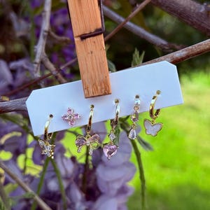 Può includere: Un set di sei orecchini dorati esposti su una carta bianca, con vari design. Gli orecchini includono forme di farfalla, fiocco, cuore e floreali, adornati con pietre preziose rosa e trasparenti. Lo sfondo include fiori viola e fogliame verde.