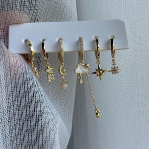 Può includere: Un set di sei orecchini a cerchio dorati con ciondoli celesti, tra cui stelle, lune, nuvole e un pianeta. Ogni orecchino presenta piccoli abbellimenti scintillanti. Gli orecchini sono esposti su una carta bianca su uno sfondo morbido e neutro.