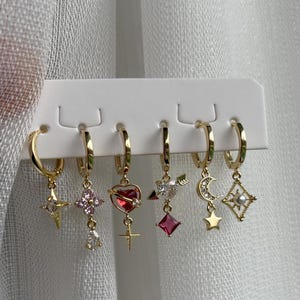 May include: A set of six gold-colored hoop earrings with various charms. Charms include stars, hearts, crosses, moons, and geometric shapes, some with pink and red accents. The earrings are displayed on a white card.