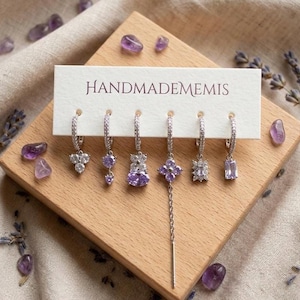 Może przedstawiać: Zestaw sześciu srebrzystych kolczyków typu koła z fioletowymi i przezroczystymi kryształkami, umieszczonych na białej karcie z napisem HANDMADEMEMIS. Kolczyki mają różne wzory, w tym kształty kwiatów, wiszące łańcuszki i prostokątne kamienie.