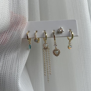 Op de afbeelding: Een set van zes goudkleurige oorbellen, gepresenteerd op een witte kaart. De oorbellen hebben verschillende ontwerpen, waaronder druppelvormige edelstenen, opalescerende stenen, hartvormige hangers en parelstuds. De achtergrond is witte stof.