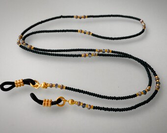 Könnte beinhalten: Schwarze Perlen-Brillenschnur mit goldenen und schillernden Perlen. Die Schnur hat zwei schwarze Gummieenden mit goldenen Akzenten.
