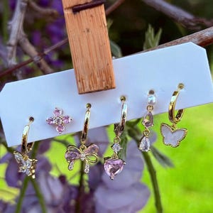 Può includere: Un set di cinque orecchini color oro su un cartoncino bianco. La collezione include un cerchio a farfalla, un perno floreale rosa, un cerchio a fiocco con pietre rosa, un orecchino a goccia a forma di cuore e un cerchio con ciondolo a farfalla.