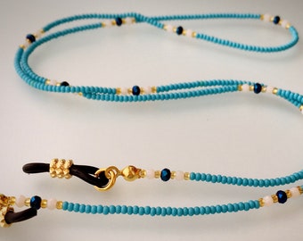 May include: A turquoise and white beaded eyeglass chain with gold-toned metal clasps. The chain is made of small, round beads and features dark blue accent beads.