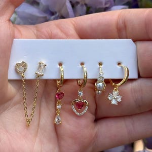 Peut inclure: Un ensemble de six boucles d'oreilles dorées sur un support blanc. La collection comprend une boucle d'oreille à double clou reliée par une chaîne, une boucle pendante avec un cœur rouge, un anneau à breloque cœur, une perle et une fleur.