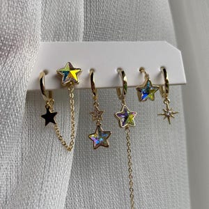 Op de afbeelding: Een verzameling goudkleurige sterrenoorbellen, gepresenteerd op een witte kaart. De oorbellen hebben verschillende ontwerpen, waaronder zwarte sterren, iriserende sterren en sterrenbedels. Sommige oorbellen hebben kettingdetails.