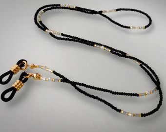 Könnte beinhalten: Schwarze Perlen-Brillenschnur mit goldenen Akzenten und weißen Perlen. Die Schnur hat schwarze Gummieinsätze zum Halten der Brille.