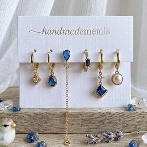 May include: A set of five gold-toned hoop earrings displayed on a white card labeled handmadememis. The earrings feature various charms including a clear square stone, a blue teardrop, a long chain, a blue baguette, a blue diamond, and a pearl.