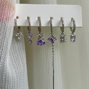 Könnte beinhalten: Eine Auswahl von sechs silberfarbenen Creolen mit verschiedenen Edelstein-Designs. Die Ohrringe haben klare und violette Edelsteine in Blumen-, Tropfen- und geometrischen Formen. Die Ohrringe werden auf einem weißen Halter präsentiert.