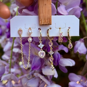 May include: A collection of gold-toned earrings with pink and white accents. The earrings feature various designs, including teardrops, hearts, and floral shapes. Some earrings have chain details. The earrings are displayed against a backdrop of purple flowers.
