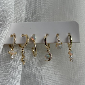 Könnte beinhalten: Ein Set aus sechs goldfarbenen Creolen. Jede Ohrring hat ein einzigartiges Design, einschließlich opalisierender Steine, hängender Charms und funkelnder Akzente. Die Ohrringe werden auf einer weißen Karte vor einem strukturierten weißen Hintergrund präsentiert.