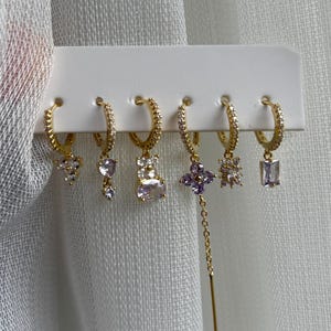 May include: A set of six gold-toned hoop earrings with various dangling charms. The charms include clear and purple crystals in different shapes, such as hearts, flowers, and rectangles. The earrings are displayed on a white card.