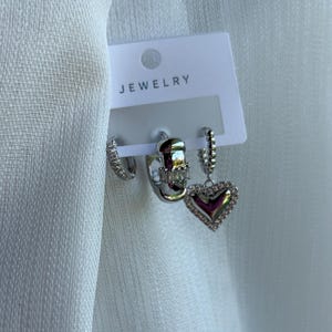 May include: A set of three earrings on a white card labeled "JEWELRY". The earrings include a small hoop with clear stones, a silver hoop with a rectangular design, and a heart-shaped earring with clear stones.
