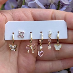 May include: A set of five gold-tone earrings on a white display card, including a butterfly stud with pink stones, a butterfly hoop, a bow hoop, a heart drop hoop with pink stones, and a pearl and teardrop hoop with a white butterfly charm.