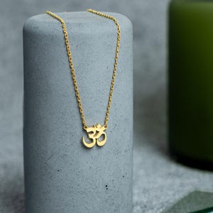 Puede incluir: Un collar dorado con el símbolo Om, exhibido sobre un soporte cilíndrico gris. La fina cadena del collar es el punto focal. Un jarrón verde con flores moradas y fragmentos de vidrio se encuentran en el fondo.