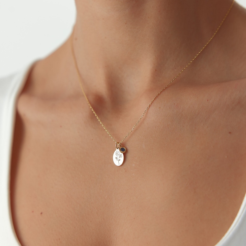 May include: A gold-toned necklace featuring a delicate chain and a small oval pendant engraved with a floral design. Beside the oval pendant hangs a tiny, circular dark blue gemstone charm, both suspended from the thin metallic link chain.