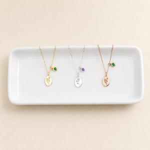 May include: Three necklaces with oval pendants featuring engraved floral designs. The first is gold-toned with a green stone, the second is silver-toned with a purple stone, and the third is rose gold-toned with a green stone, all on thin chains.