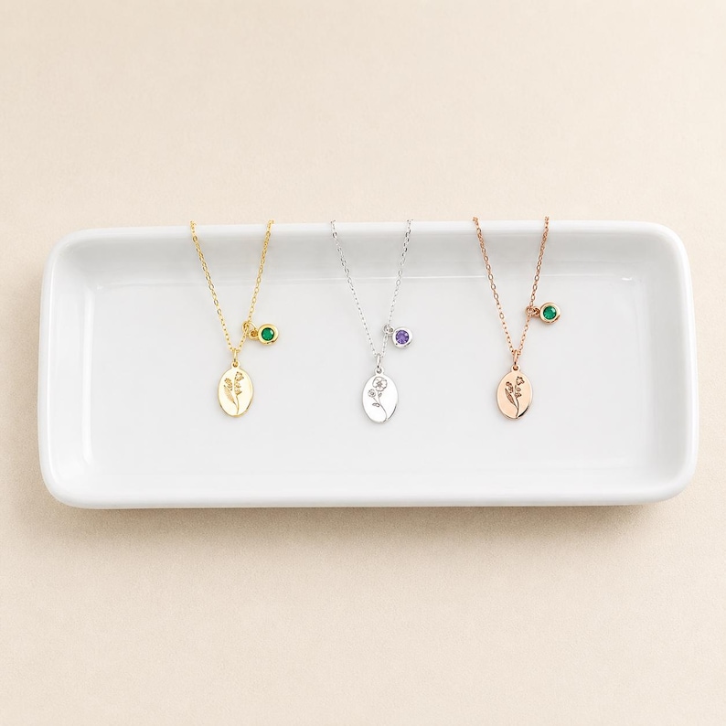 May include: Three necklaces with oval pendants featuring engraved floral designs. The first is gold-toned with a green stone, the second is silver-toned with a purple stone, and the third is rose gold-toned with a green stone, all on thin chains.