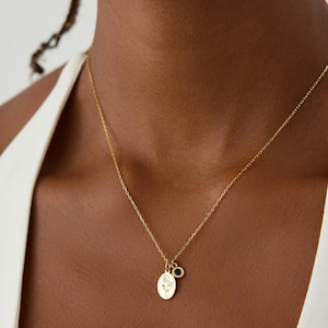 May include: A gold chain necklace featuring two charms: an oval pendant engraved with a delicate line-art floral design and a small, circular charm set with a single, dark blue stone. The necklace is worn around the neck with a white garment.