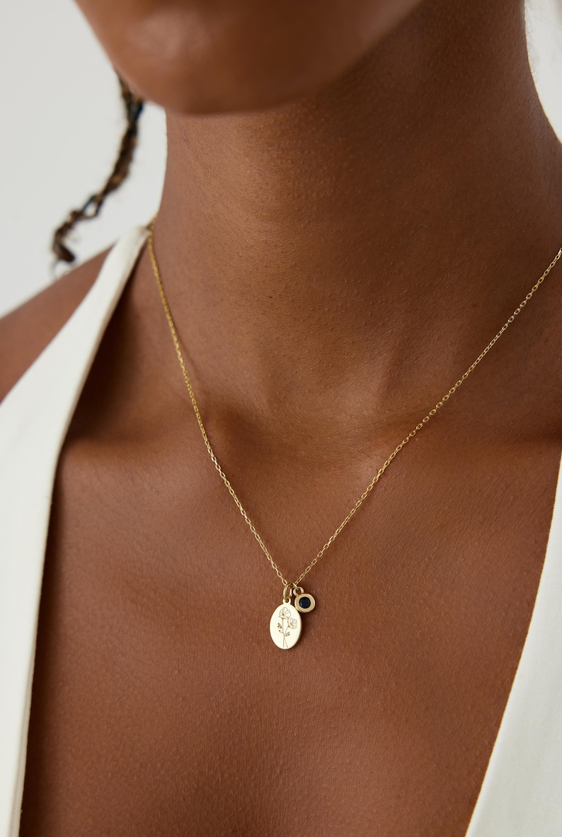 May include: A gold chain necklace featuring two charms: an oval pendant engraved with a delicate line-art floral design and a small, circular charm set with a single, dark blue stone. The necklace is worn around the neck with a white garment.