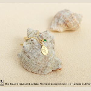 May include: A gold-toned necklace featuring a small oval pendant engraved with a floral design and a separate circular charm set with a single green stone. The delicate chain is draped over a textured, light-colored seashell.