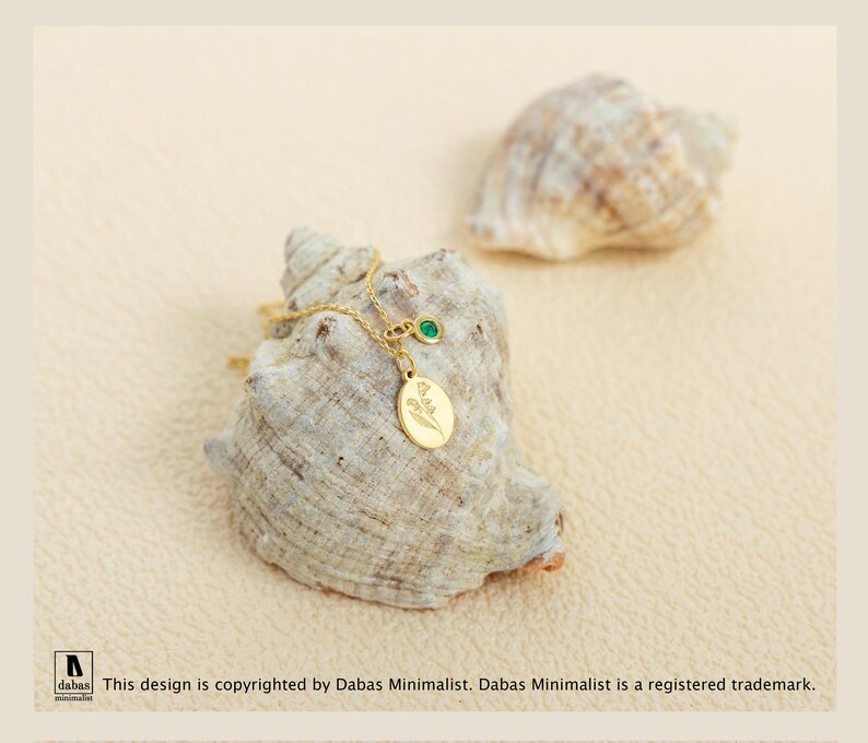 May include: A gold-toned necklace featuring a small oval pendant engraved with a floral design and a separate circular charm set with a single green stone. The delicate chain is draped over a textured, light-colored seashell.