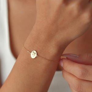 Puede incluir: Una pulsera de cadena en tono dorado con un peque&ntilde;o colgante circular. El colgante presenta un grabado con una delicada ilustraci&oacute;n de l&iacute;neas finas de dos flores con tallos y hojas, centradas en la superficie met&aacute;lica lisa y pulida.