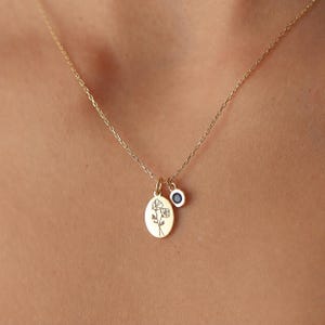 May include: A gold chain necklace featuring an oval pendant engraved with a delicate floral line drawing and a small circular charm set with a dark blue stone. The necklace rests against the skin, showcasing the polished gold metal finish.