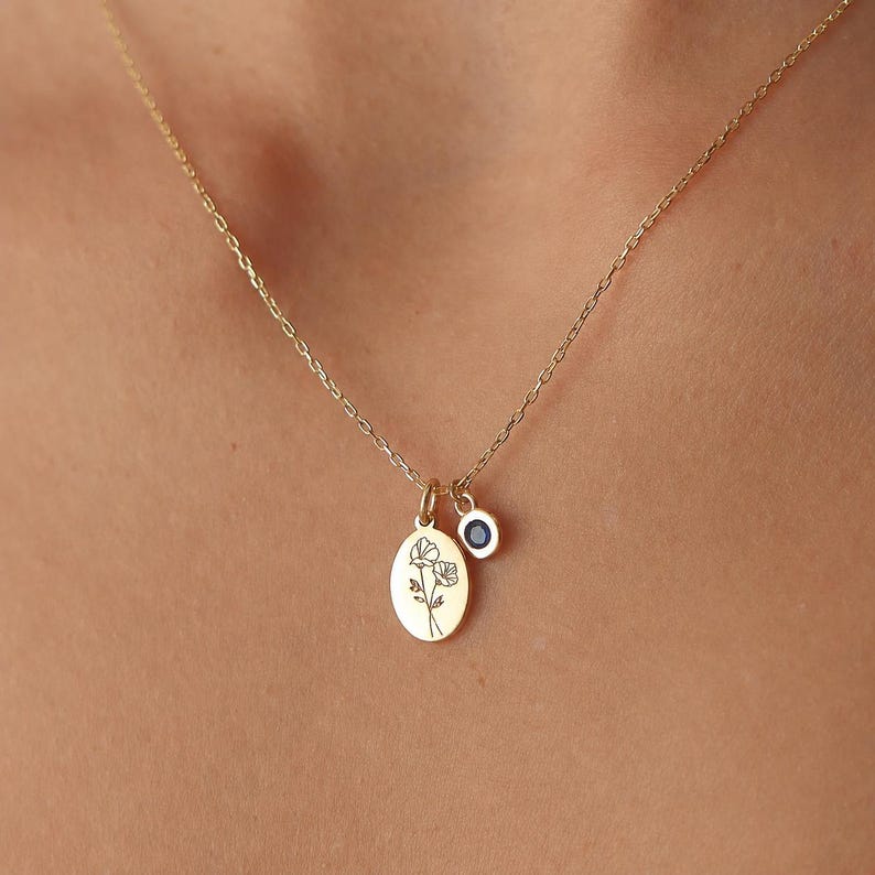 May include: A gold chain necklace featuring two pendants: an oval-shaped gold charm engraved with a delicate line drawing of two flowers and a smaller circular gold charm set with a single dark blue gemstone, suspended from a thin gold link chain.