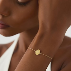 Puede incluir: Una pulsera de cadena en tono dorado con un peque&ntilde;o colgante circular. El colgante est&aacute; grabado con una delicada ilustraci&oacute;n floral de l&iacute;neas finas de una sola flor. La pulsera se asegura con una cadena fina de metal pulido color oro.