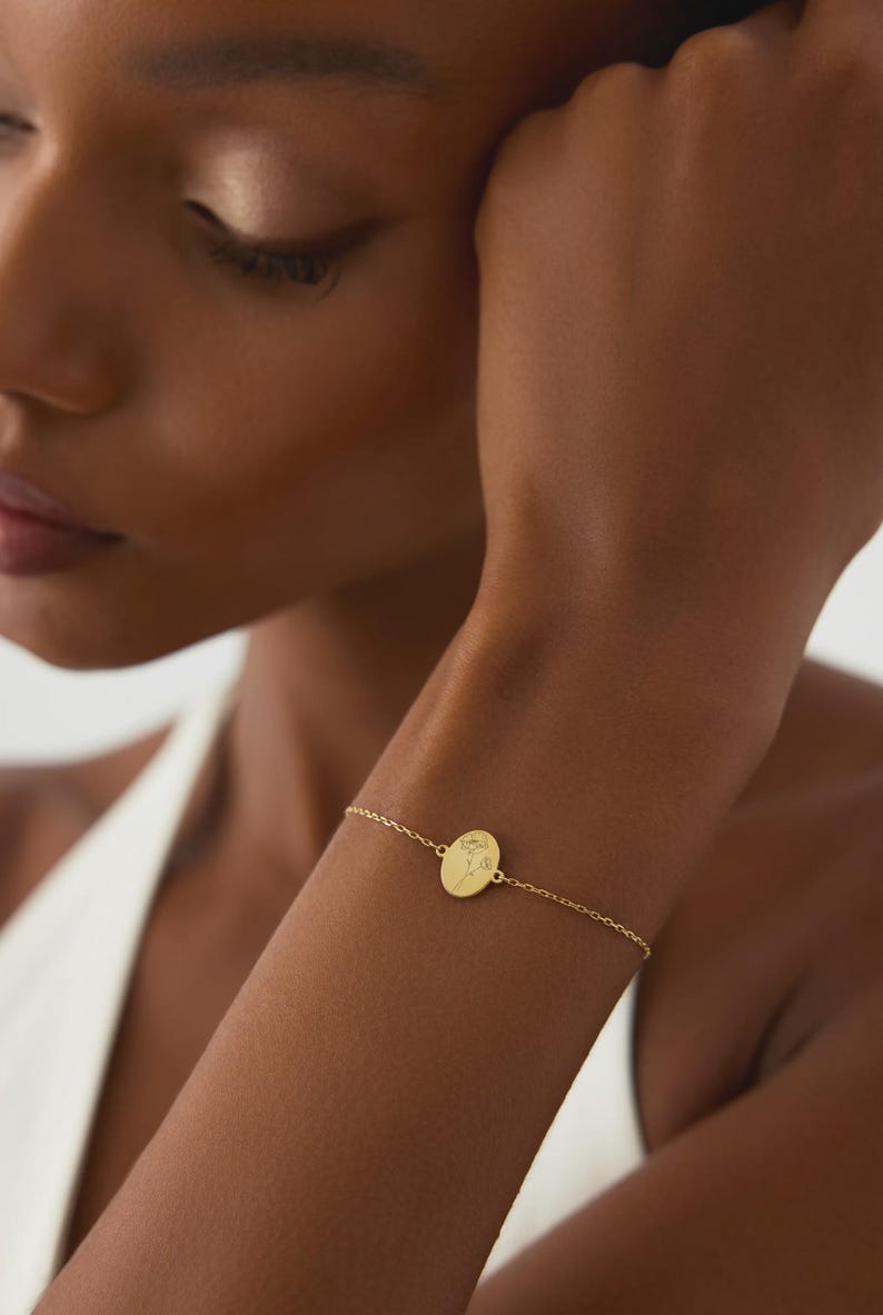 Puede incluir: Una pulsera de cadena en tono dorado con un peque&ntilde;o colgante circular. El colgante est&aacute; grabado con una delicada ilustraci&oacute;n floral de l&iacute;neas finas de una sola flor. La pulsera se asegura con una cadena fina de metal pulido color oro.