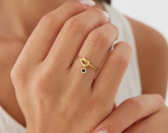 May include: A gold-toned ring featuring a delicate knot design on the band. A small, circular charm with a dark blue stone is suspended from the knot, creating a subtle dangling effect on this thin, polished metal finger ring accessory.