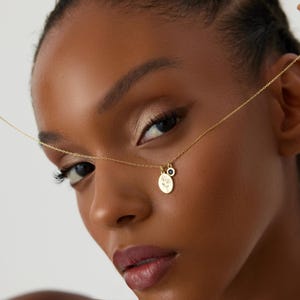 May include: A gold-tone chain necklace featuring two small pendants: one oval-shaped charm with an engraved floral design and a second, smaller circular charm with a dark blue stone inlay. The delicate metal chain connects to both charms.