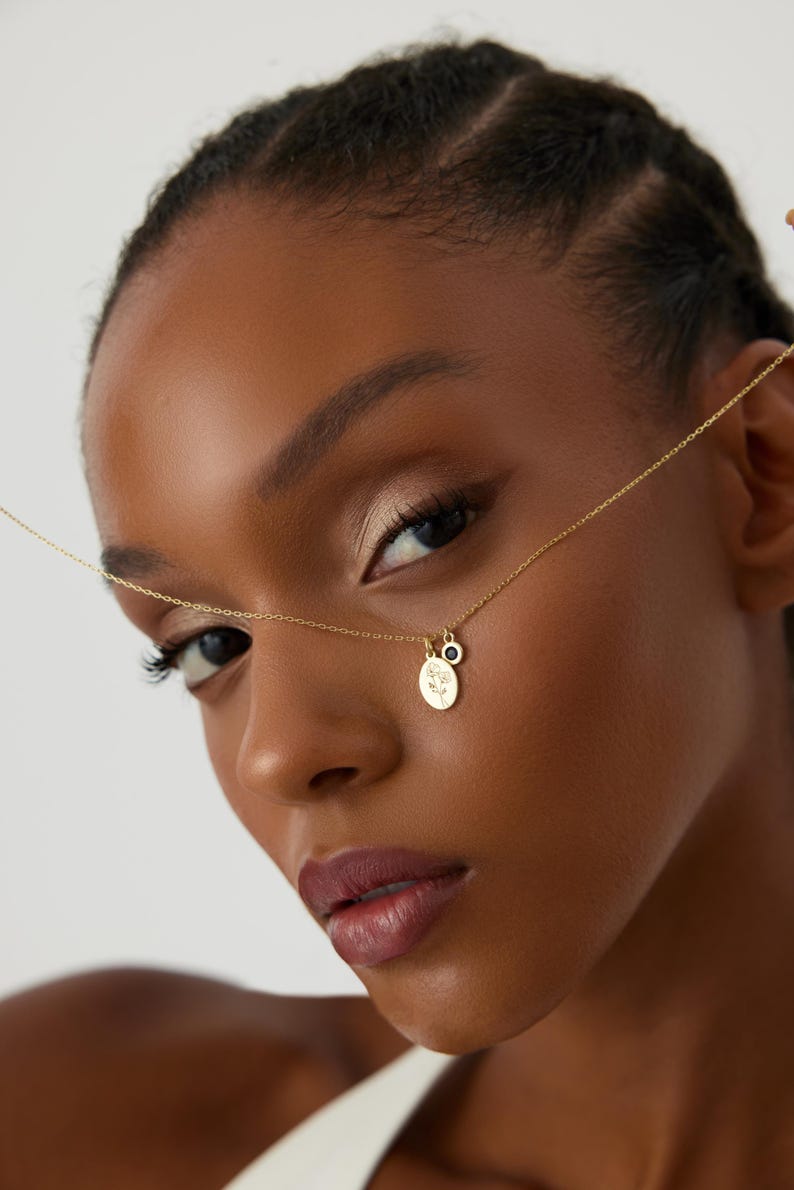 May include: A gold-tone chain necklace featuring two small pendants: one oval-shaped charm with an engraved floral design and a second, smaller circular charm with a dark blue stone inlay. The delicate metal chain connects to both charms.